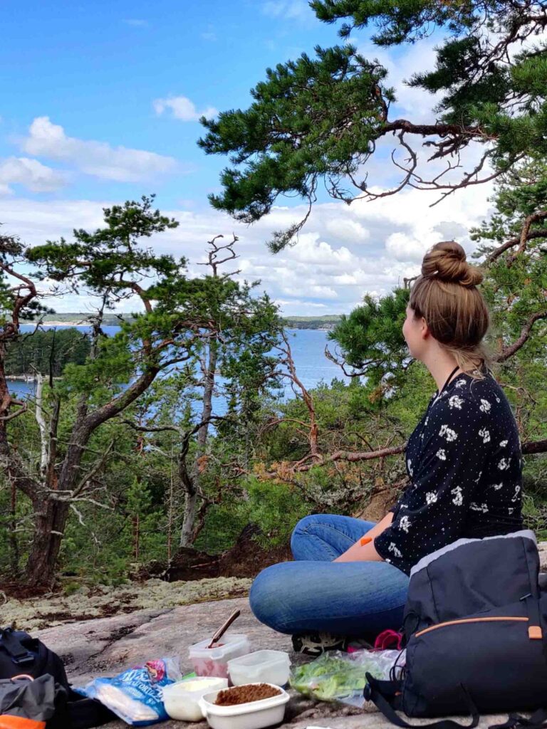 Kallio, jossa istuu nuori nainen. Taustalla mäntyjä ja merimaisema, etualalla eväitä.