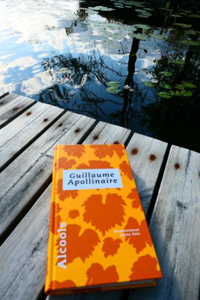 Kuvassa oranssinvärinen kirja, kannessa lehtikuvituksia. Teksti Guillaume Apollinaire mustalla värillä valkoisella taustalla. Alcools valkoisella tekstillä kannen vasemmalla reunalla.  Kirja on laiturin päällä ja taustalla näkyy lumpeenlehtien peittämä järvi. 